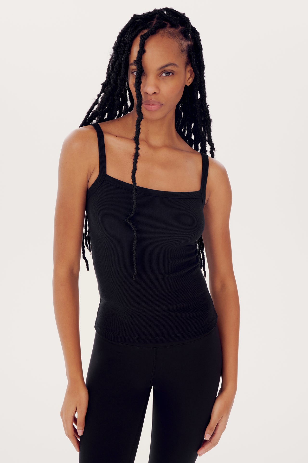 A woman in a black Romy Rib Tank by SPLITS59 and leggings, standing against a white background, looking at the camera with a neutral expression. She has long, black braided hair.