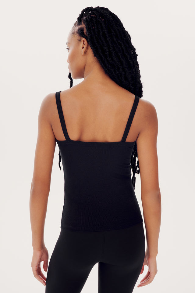 A woman with braided hair wearing a black SPLITS59 Romy Rib Tank and leggings stands with her back to the camera against a white background.
