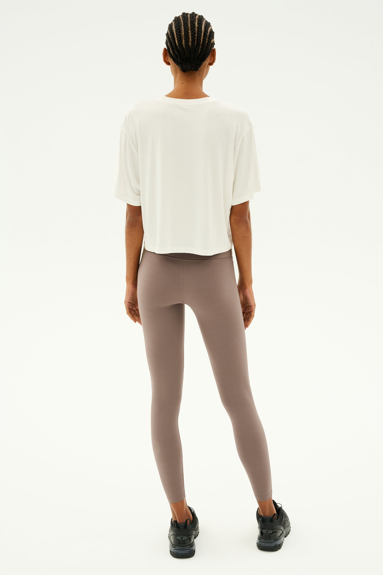 A person stands facing away, embracing the simplicity of a plain background in a Djuna Oversized Jersey Tee by SPLITS59, paired with taupe leggings and black shoes—an outfit perfect for both gym workouts and stylish comfort.