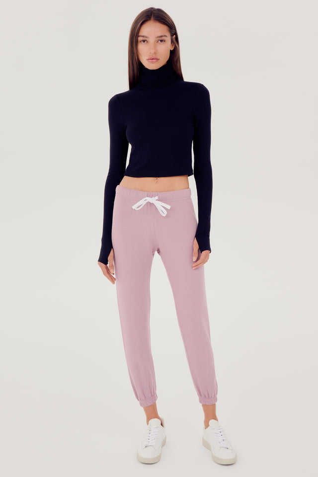 Full front view of woman with dark straight hair wearing light pink sweatpant jogger with white drawstring and black turtleneck cropped long sleeve paired with white shoes