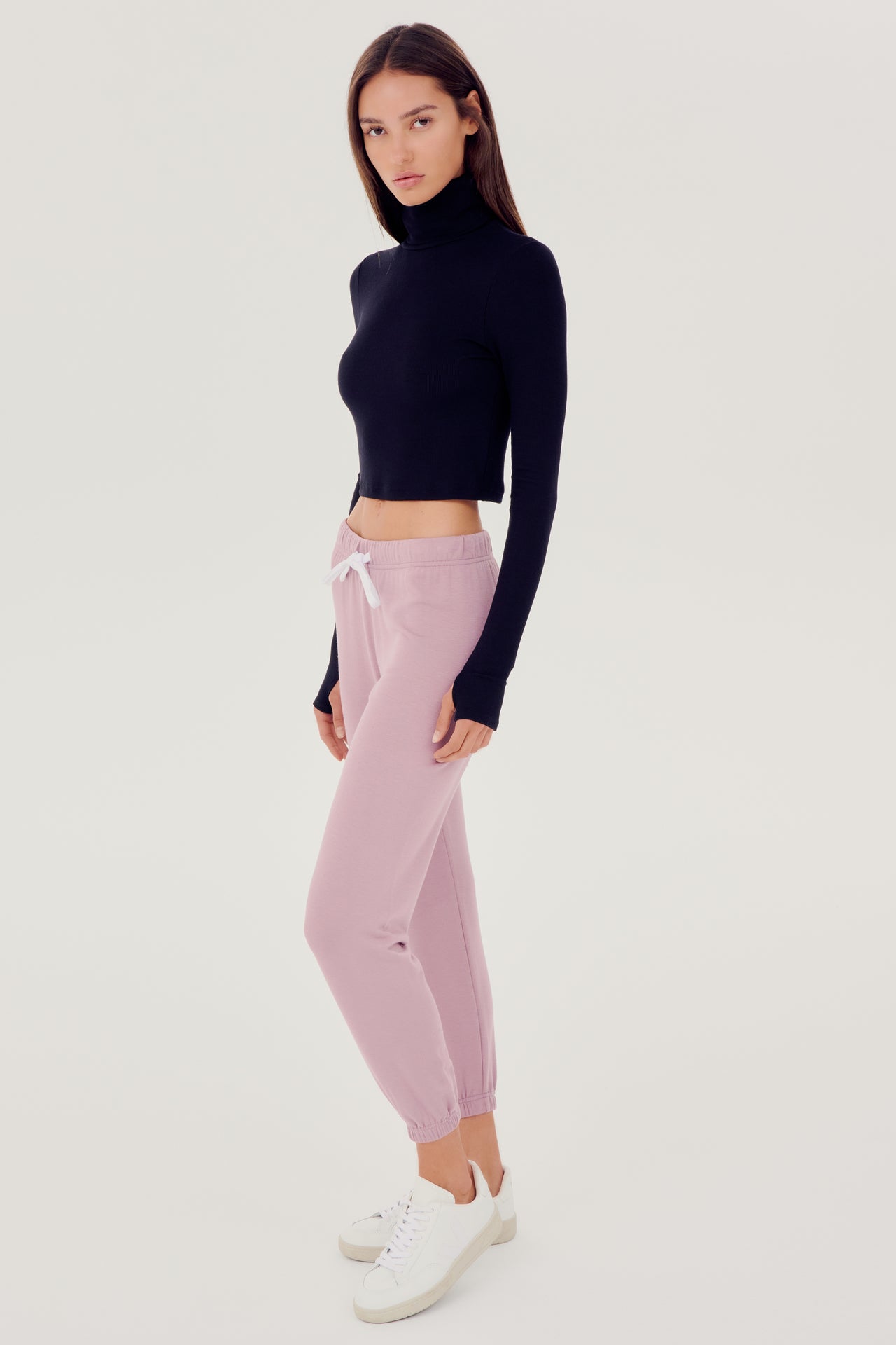 Full front side view of woman with dark straight hair wearing light pink sweatpant jogger with white drawstring and black turtleneck cropped long sleeve paired with white shoes