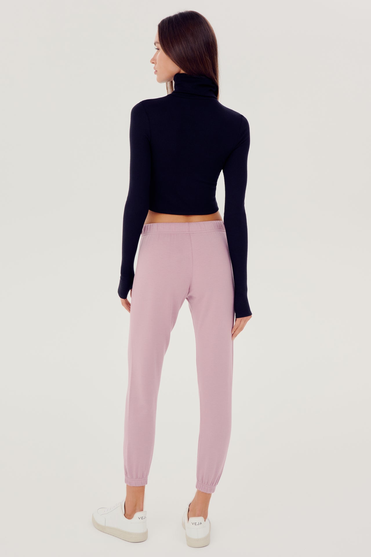 Full back view of woman with dark straight hair wearing light pink sweatpant jogger with white drawstring and black turtleneck cropped long sleeve paired with white shoes