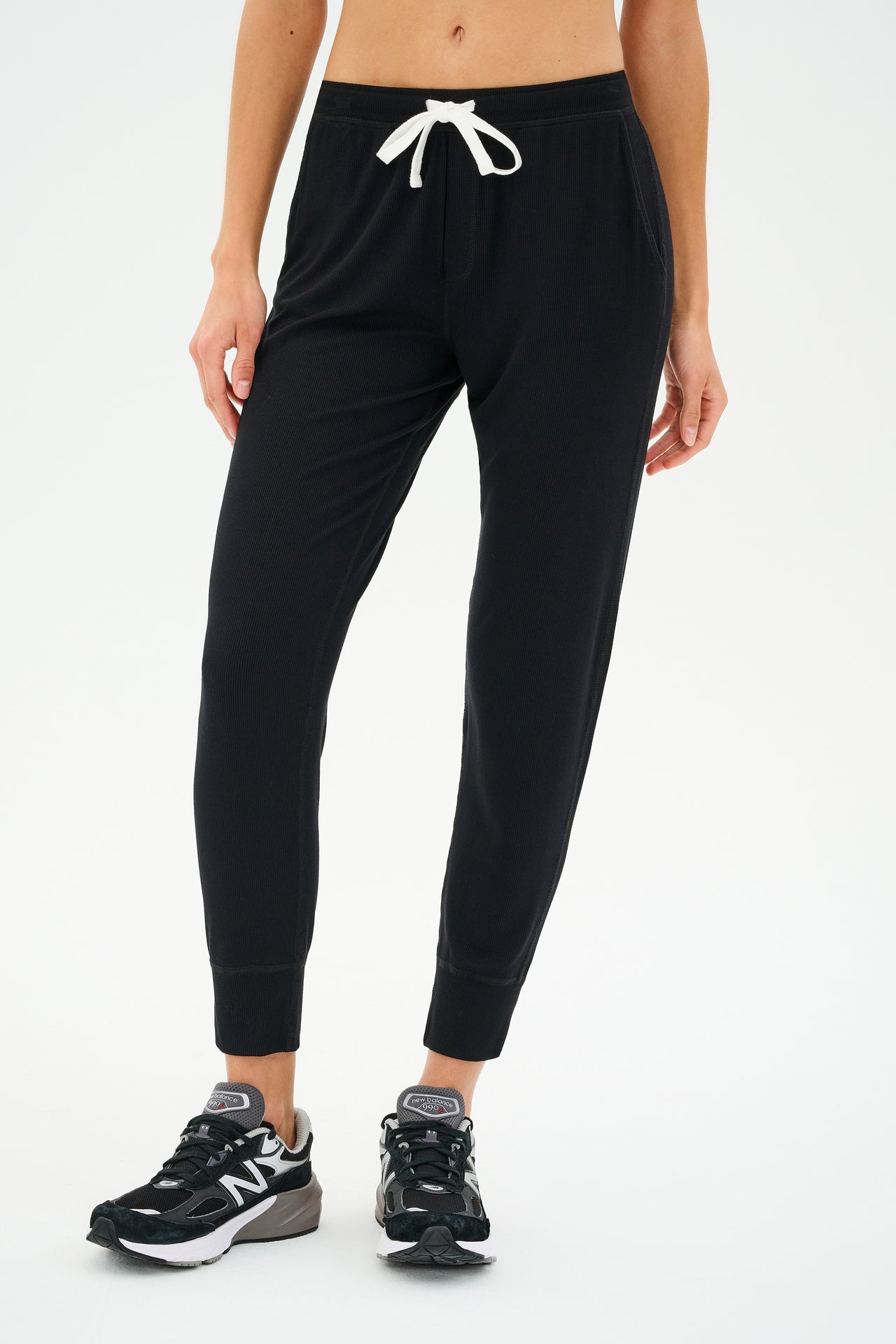 A person stands against a plain background, exuding effortless comfort in SPLITS59's Kiki Rib 7/8 Sweatpant - Black with a white drawstring, paired perfectly with sleek black sneakers.