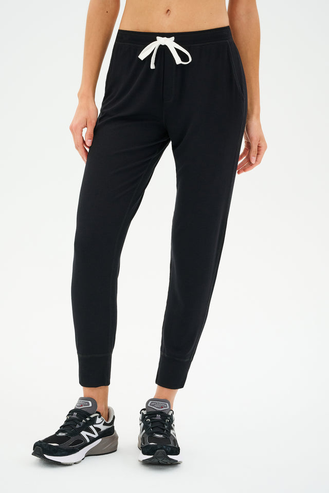 A person stands against a plain background, exuding effortless comfort in SPLITS59's Kiki Rib 7/8 Sweatpant - Black with a white drawstring, paired perfectly with sleek black sneakers.