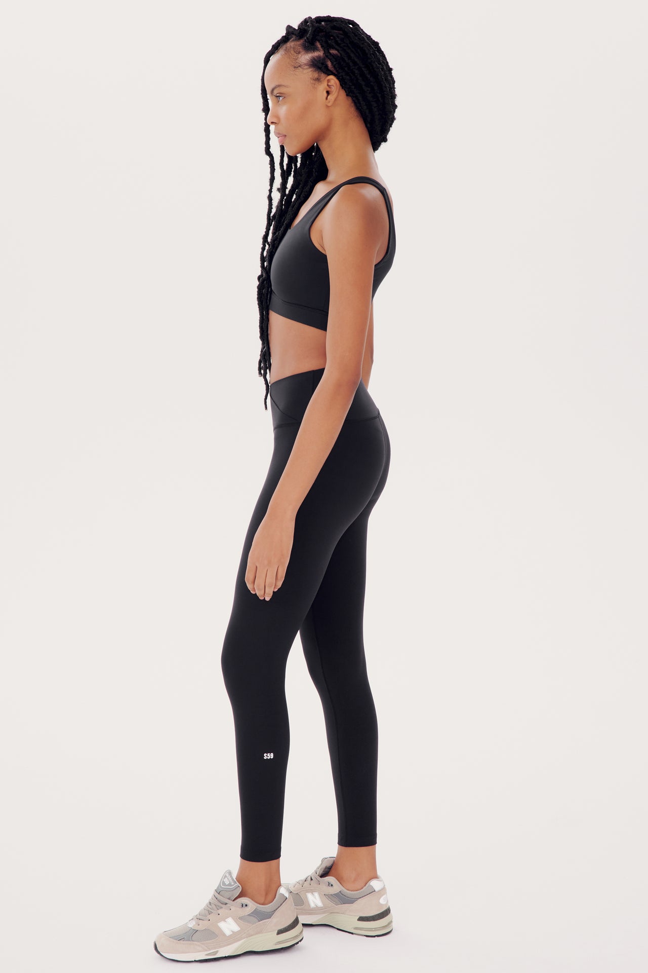 A woman in Mia Rigor 7/8 - Black sports bra and high waist leggings stands in profile, showcasing SPLITS59 activewear, with her hair in braids.