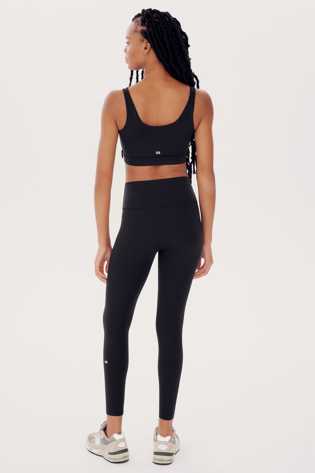 A woman in SPLITS59 black athletic wear, designed for multi-sport performance, stands with her back to the camera, showcasing the Sprint Rigor Bra - Black and her braided hair.