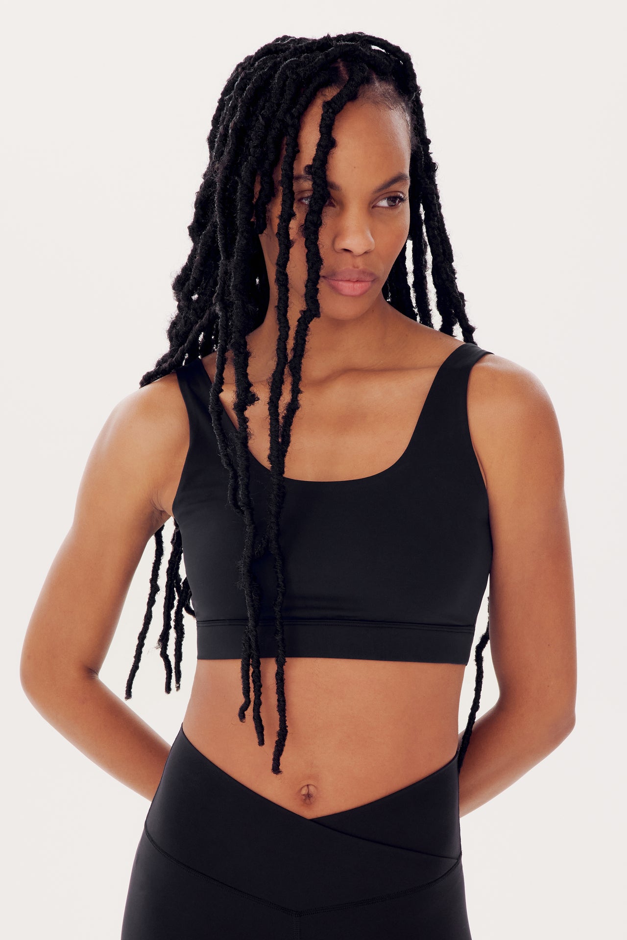 A woman with long braids wearing a SPLITS59 Sprint Rigor Bra in Black and leggings, standing against a white background, looking to the side.