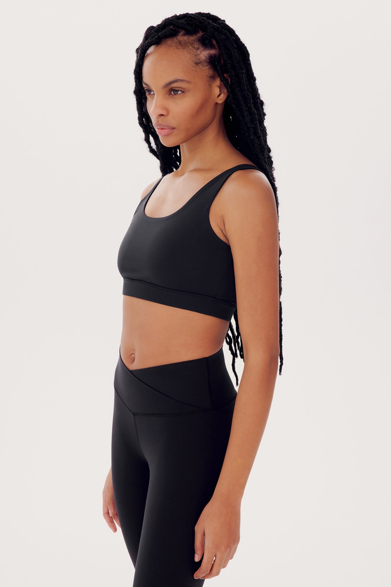 A young woman wearing a SPLITS59 Sprint Rigor Bra in Black and leggings, standing sideways for her gym workout, with long braided hair.