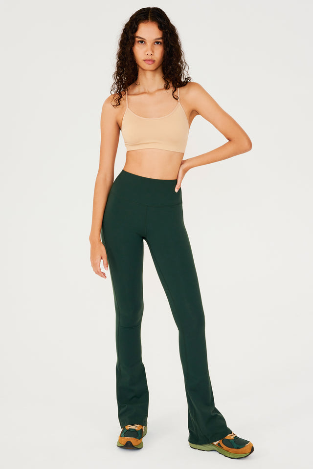 Full front view of woman with dark wavy hair wearing dark green high waist below ankle length legging with wide flared bottoms and pale warm brown bra with spaghetti straps. Paired with dark green, dark orange and light green color block shoes. 
