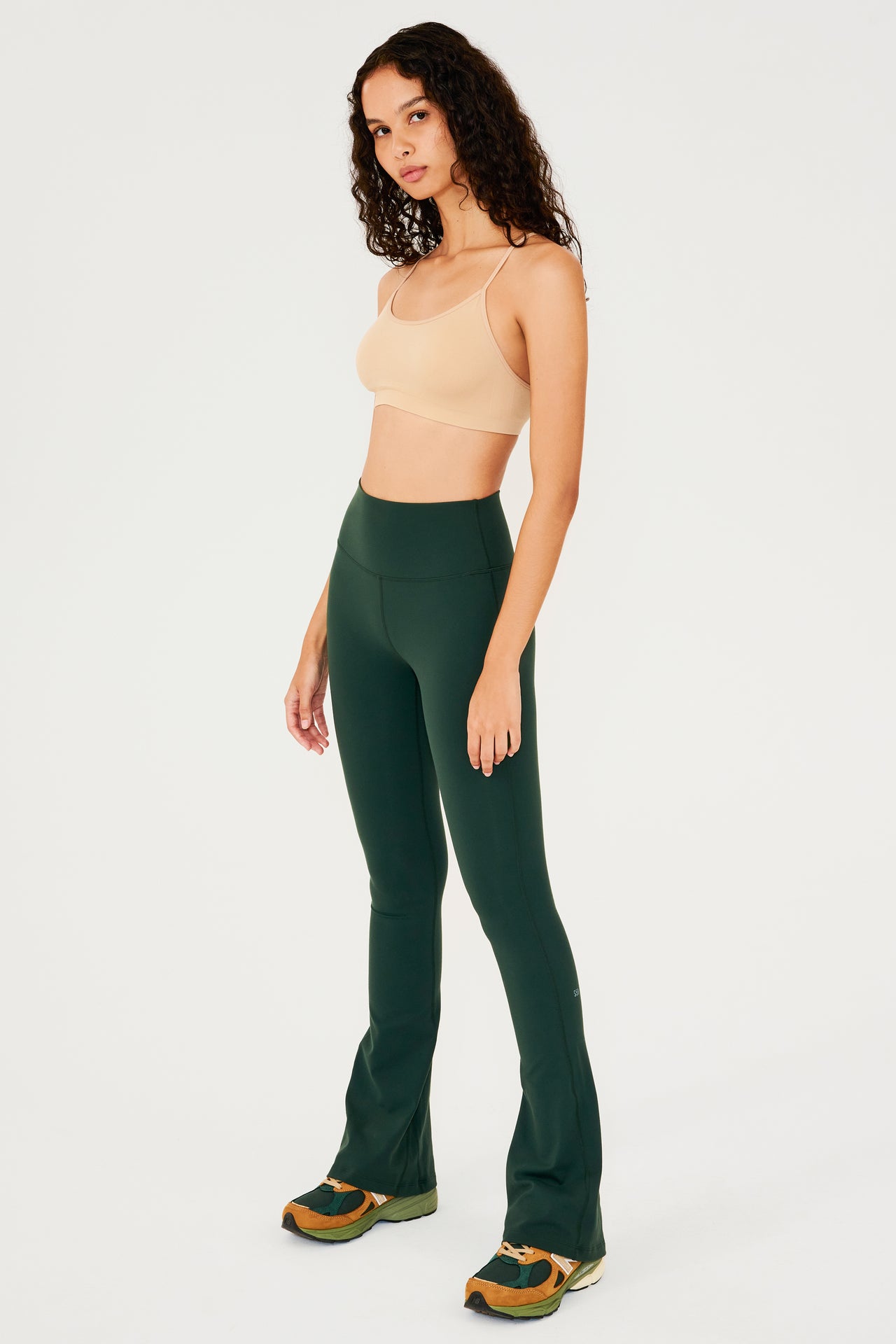 Full front side view of woman with dark wavy hair wearing dark green high waist below ankle length legging with wide flared bottoms with gray S59 logo on back of left calf and pale warm brown bra with spaghetti straps. Paired with dark green, dark orange and light green color block shoes.