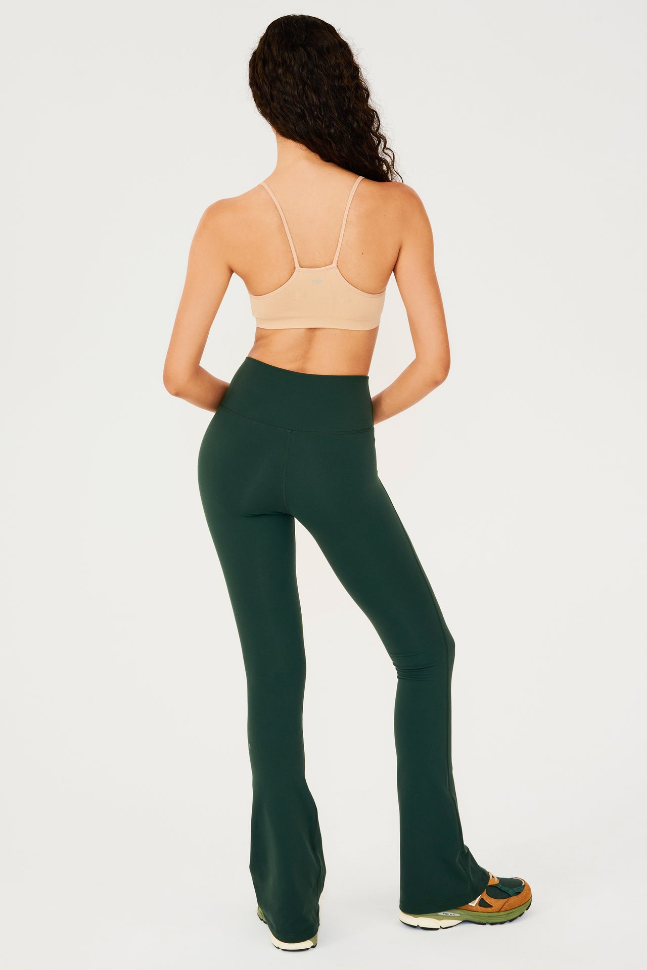 Full back view of woman with dark wavy hair wearing dark green high waist below ankle length legging with wide flared bottoms with gray S59 logo on back of left calf and pale warm brown racerback bra with spaghetti straps. Paired with dark green, dark orange and light green color block shoes.