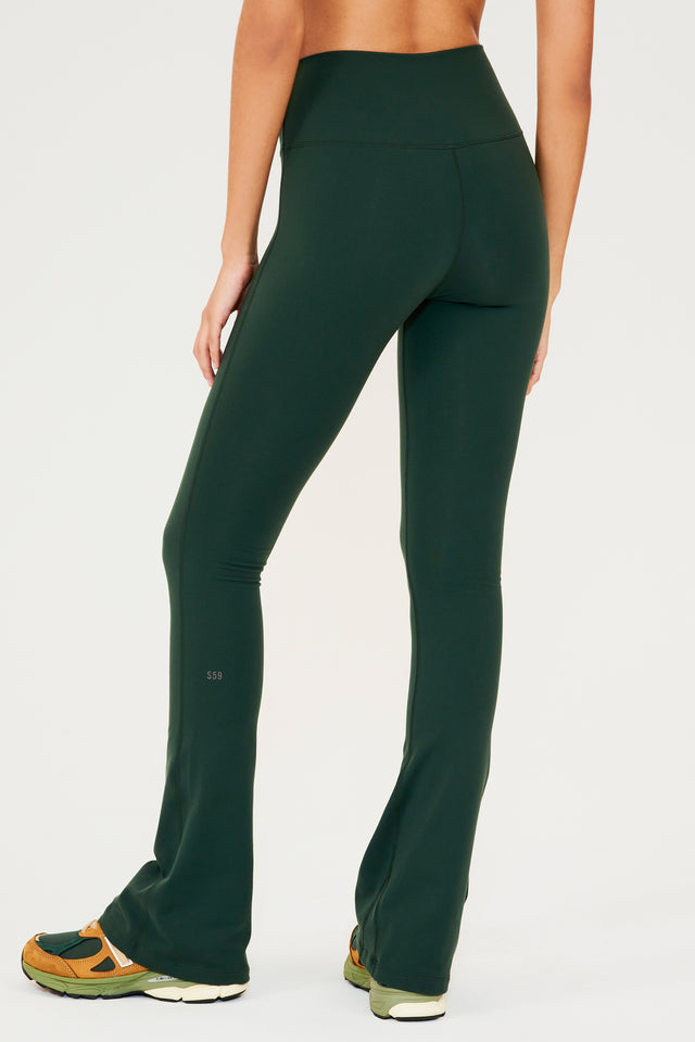 Back view of woman wearing dark green high waist below ankle length legging with wide flared bottoms with gray S59 logo on back of left calf. Paired with dark green, dark orange and light green color block shoes. 
