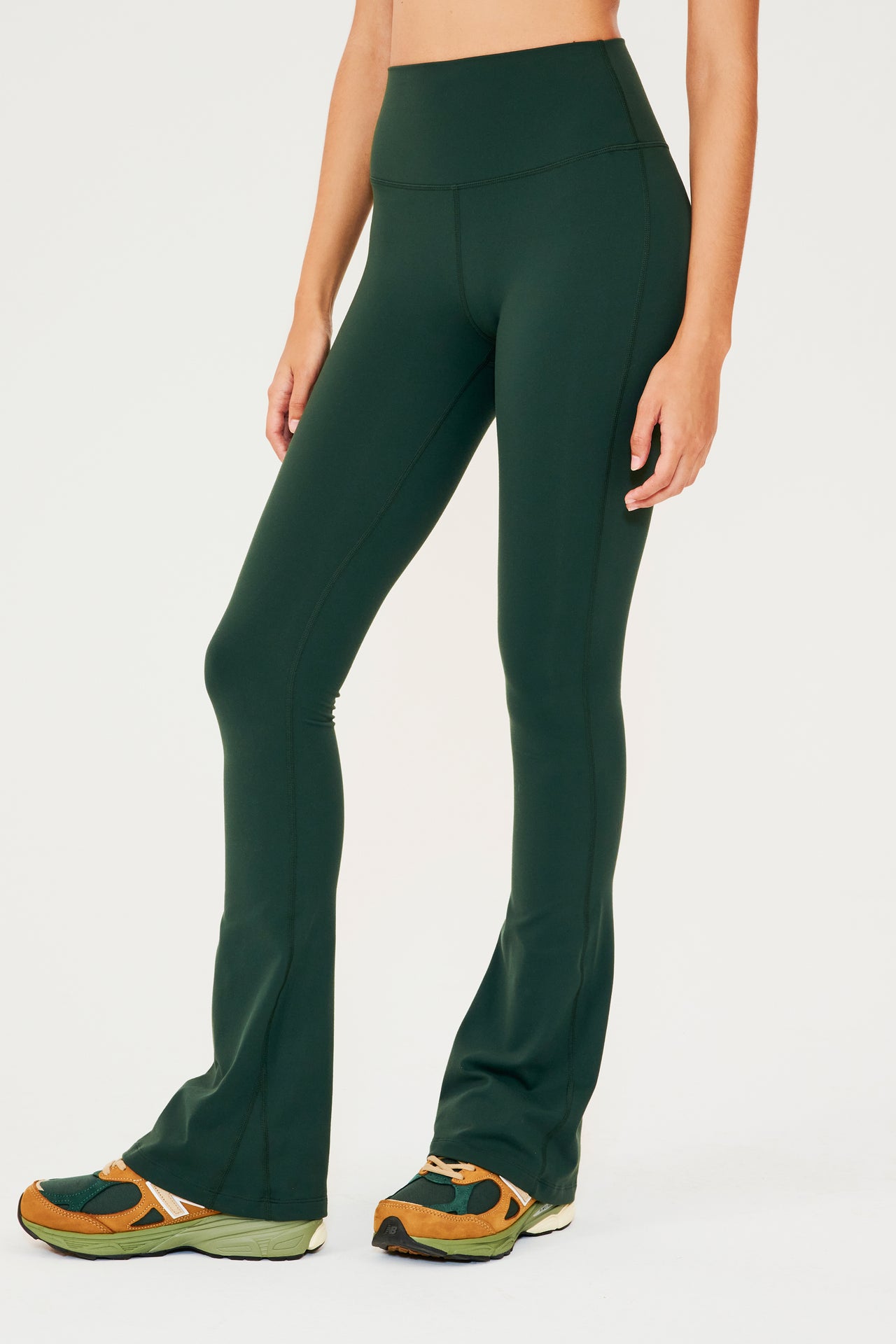 Front view of woman wearing dark green high waist below ankle length legging with wide flared bottoms. Paired with dark green, dark orange and light green color block shoes