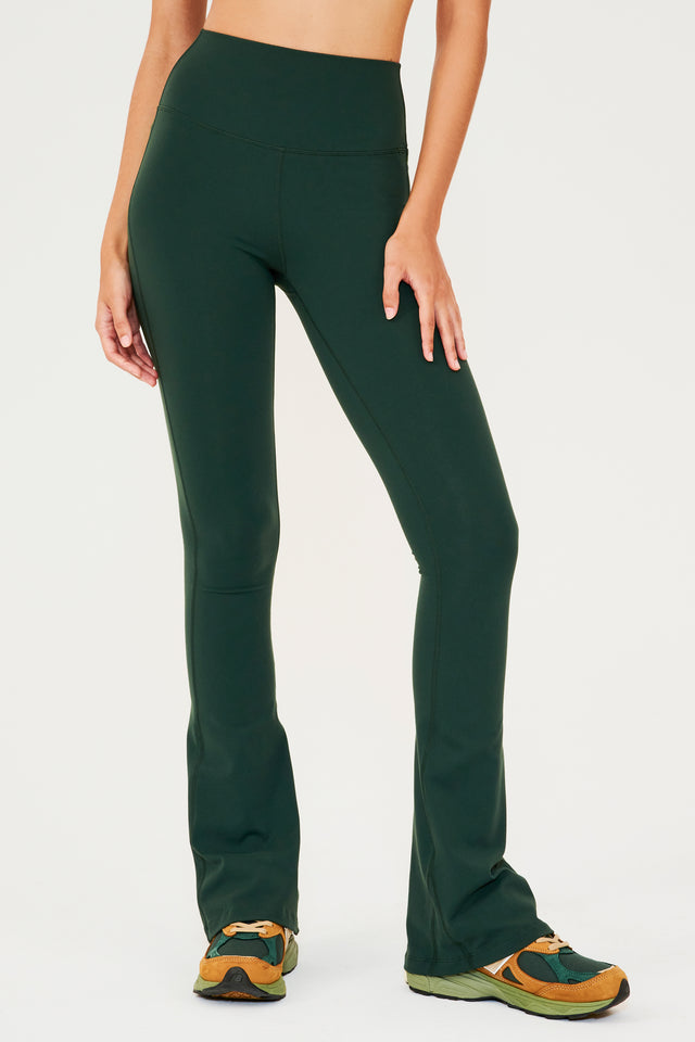Front view of woman wearing dark green high waist below ankle length legging with wide flared bottoms. Paired with dark green, dark orange and light green color block shoes. 