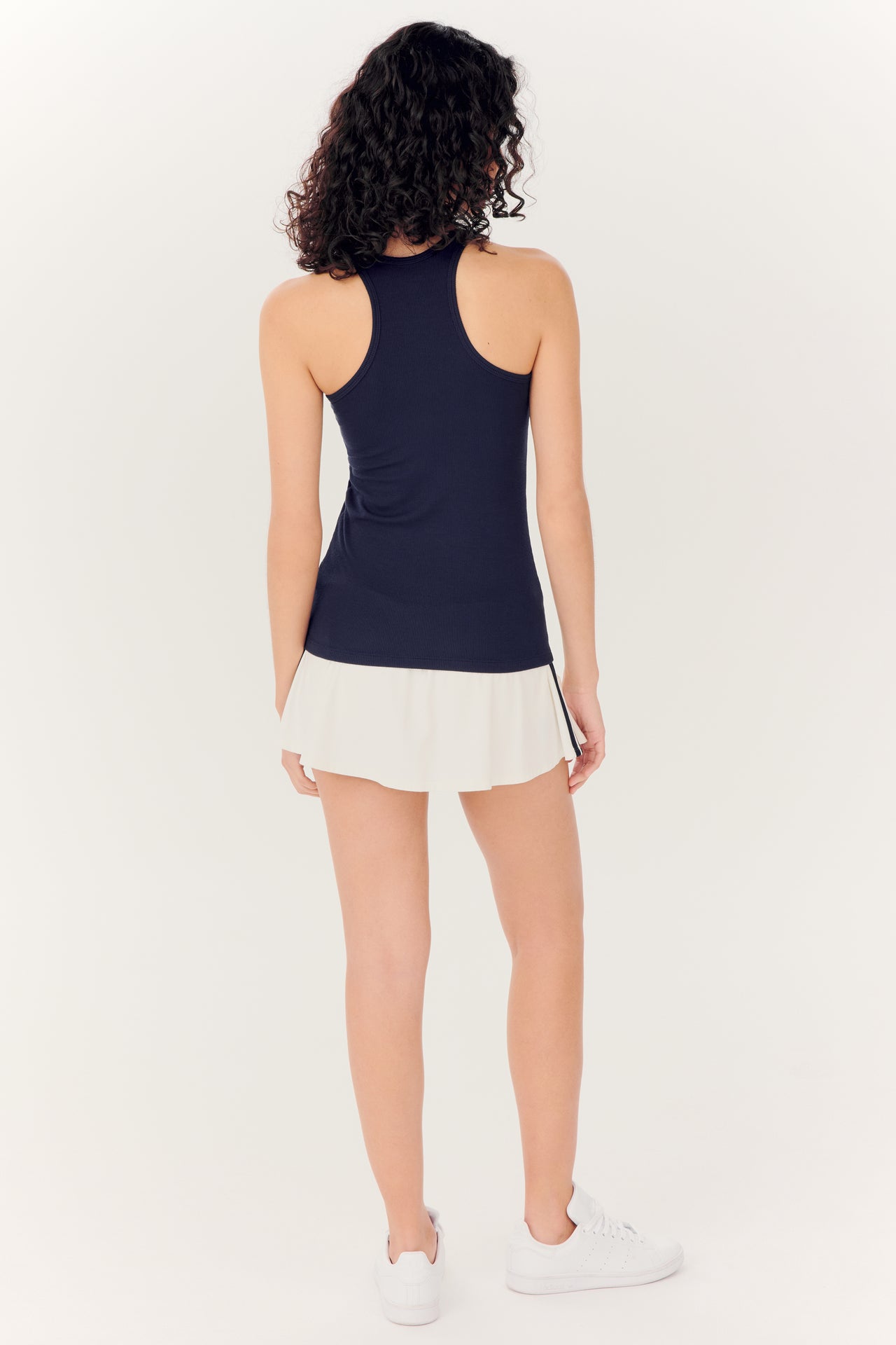 Woman from behind wearing a navy blue tank top made of SPLITS59 Airweight fabric and Ella Airweight Skort - White/Indigo with sneakers against a white background.