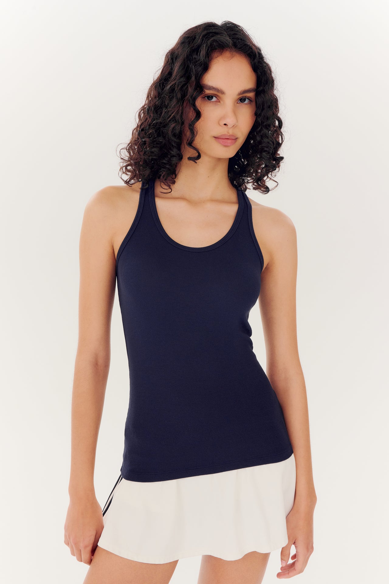 A woman with curly hair wearing a SPLITS59 Ashby Rib Tank in Indigo and a white skirt, standing against a plain white background.
