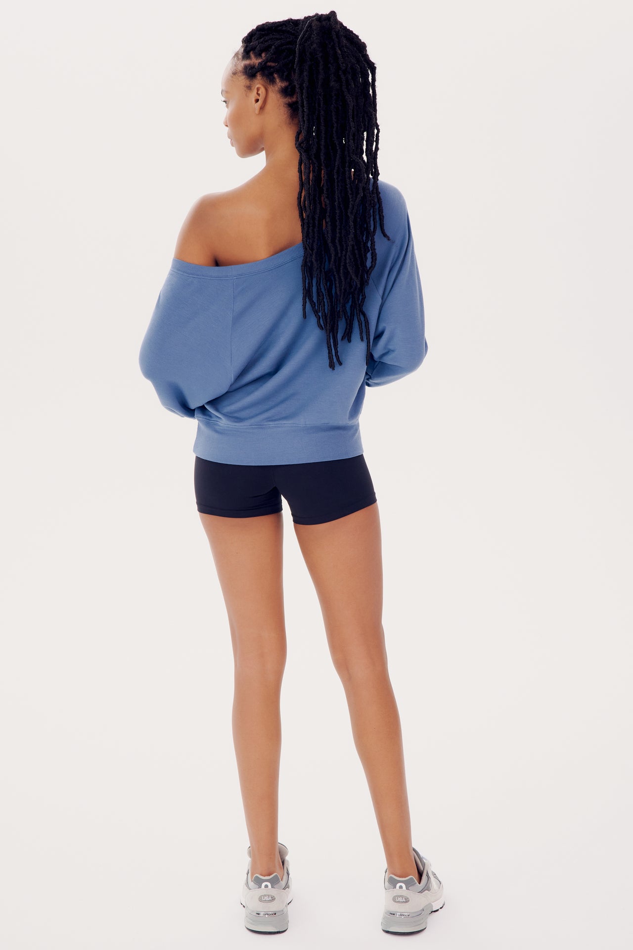 A woman in a blue SPLITS59 Indy Dolman Fleece Sweatshirt and black shorts, with braided hair, standing profile view against a white background.