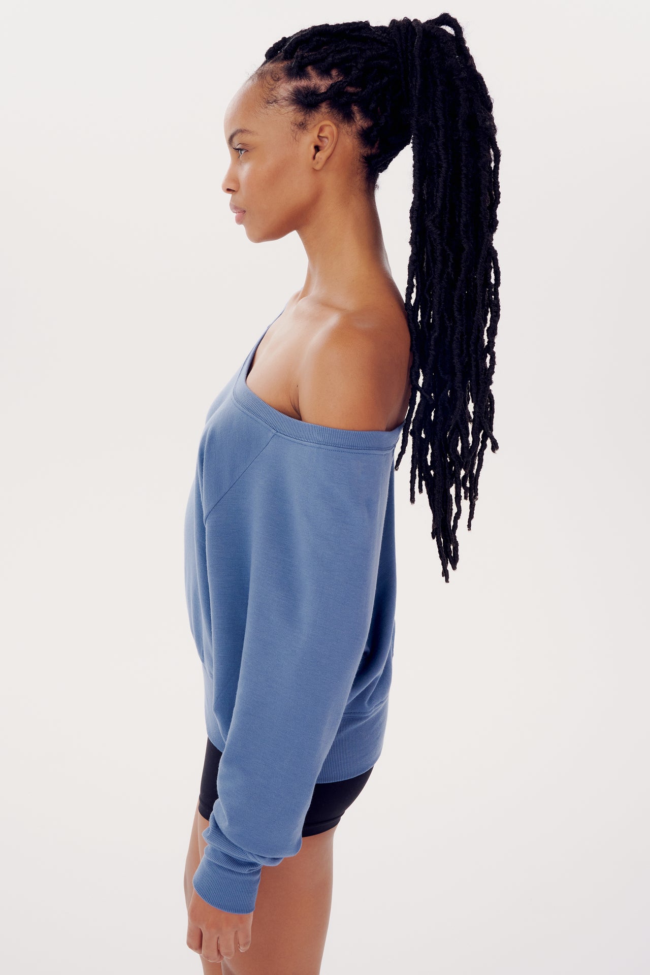A woman with long braided hair wearing a blue SPLITS59 Indy Dolman Fleece Sweatshirt in a profile view against a white background.