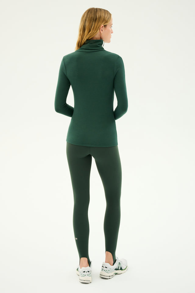 A person with long hair, wearing a supremely soft Jackson Rib Full-Length Turtleneck in Military/White from SPLITS59 and green leggings, is standing and looking to the right. The background is plain and light-colored.