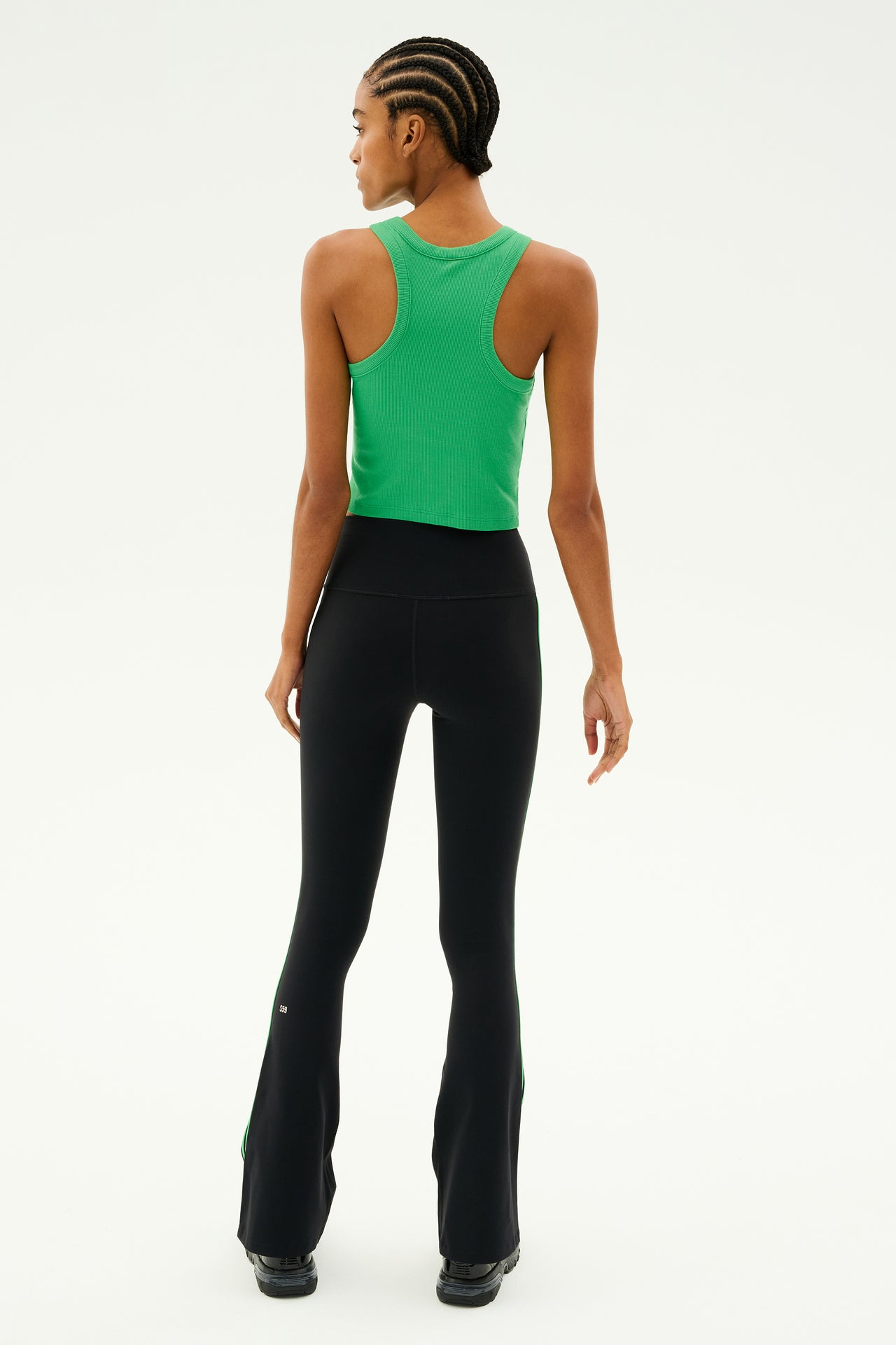 A person with braided hair wearing a green tank top and the SPLITS59 Raquel High Waist Flared Legging in Black/Grass stands with their back to the camera, prepared for yoga workouts.