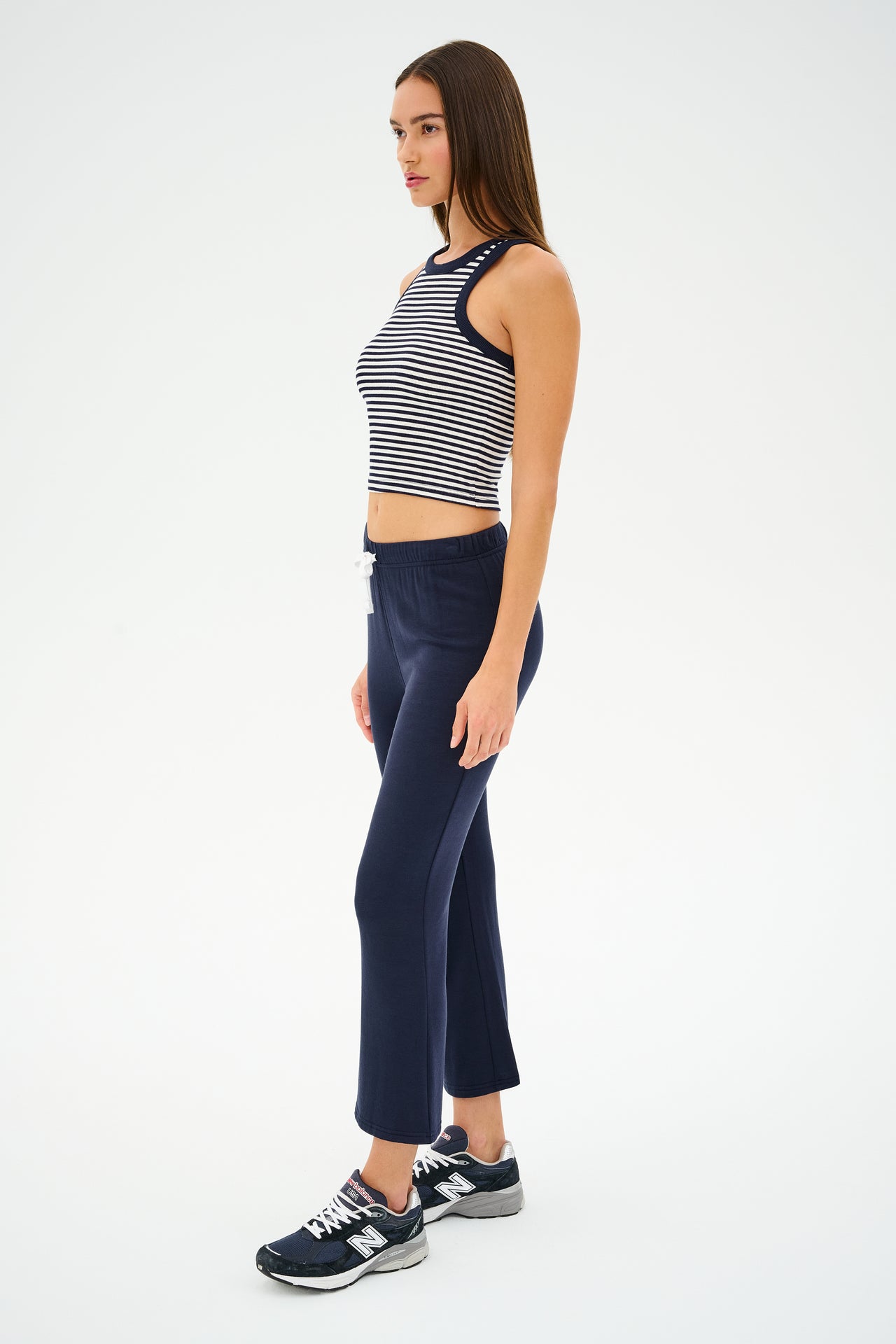 A woman in a Kiki Rib Crop Tank by SPLITS59, featuring white and indigo stripes, pairs it with navy pants and black sneakers while standing sideways against a plain background.