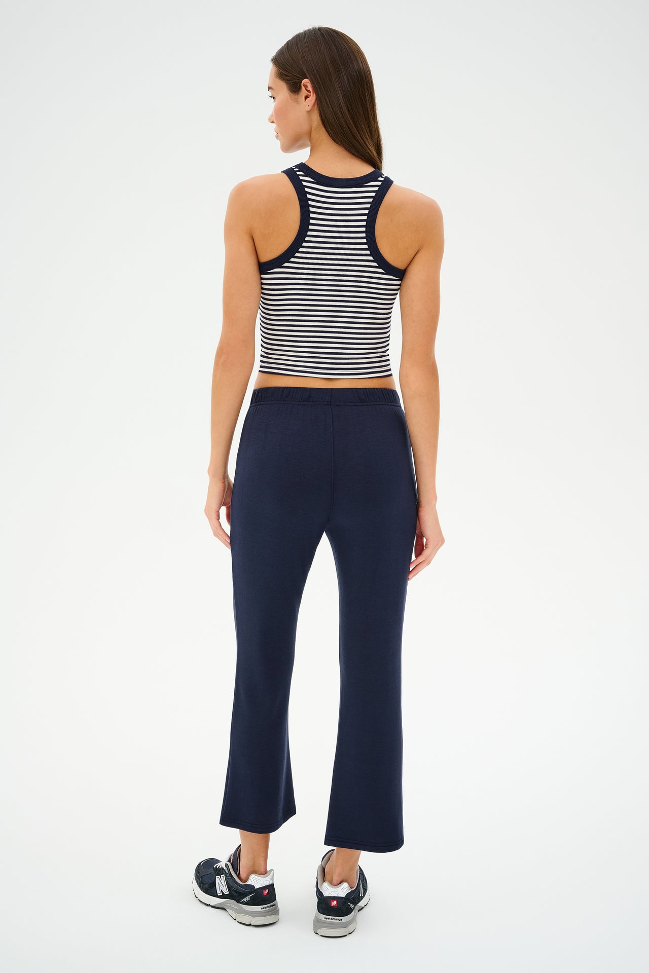 A person with long hair stands facing away, wearing the SPLITS59 Kiki Rib Crop Tank in White/Indigo Stripe, paired with dark blue pants and black sneakers.