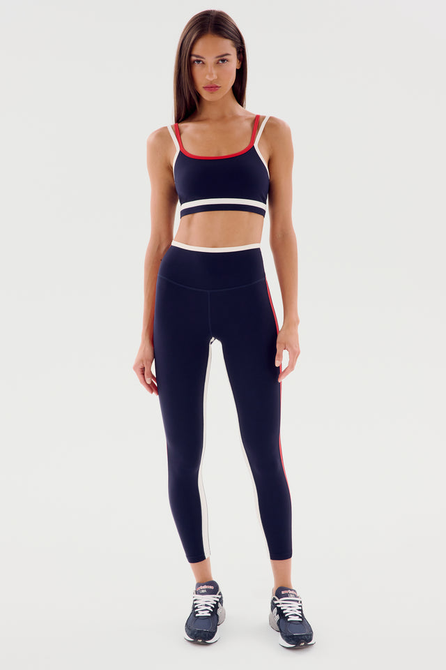 A woman wearing a navy athletic outfit with red and white accents, consisting of a SPLITS59 Sam High Waist Rigor 7/8 - Indigo/White sports bra and high-waisted leggings made from Rigor fabric, and gray running shoes, stands facing forward against a white background.