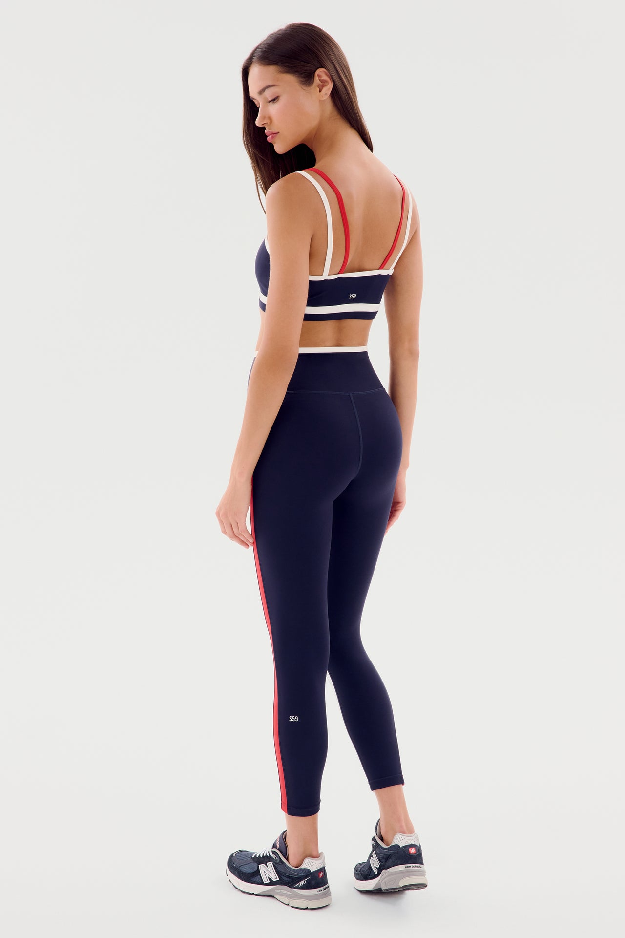 A woman is standing in profile wearing a navy blue sports bra and the Sam High Waist Rigor 7/8 - Indigo/White by SPLITS59, which feature high-waist leggings with red stripes made from rigor fabric. She completes her athletic look with black and white athletic shoes.
