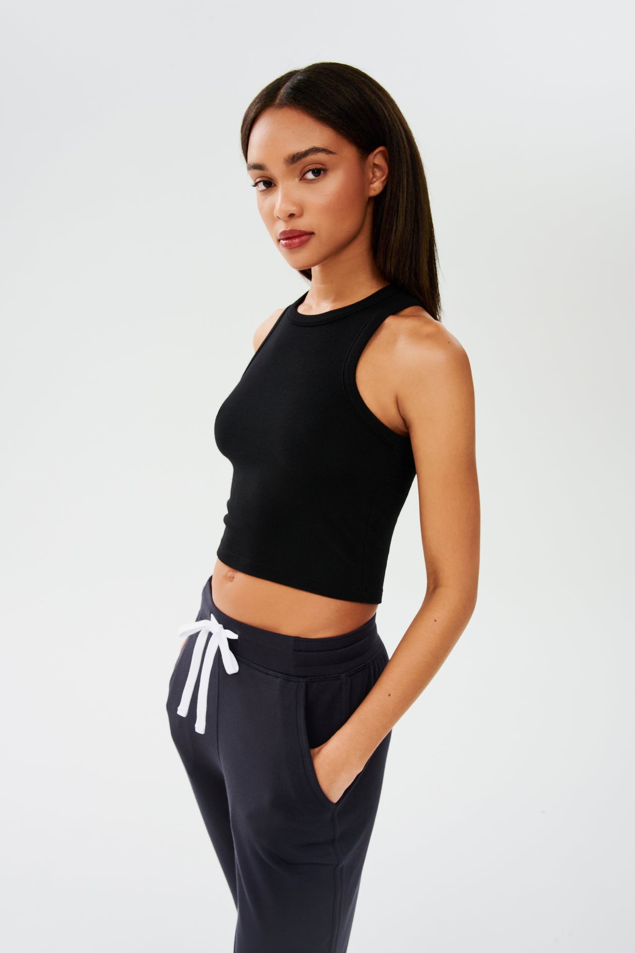The model is wearing a SPLITS59 Kiki Rib Crop Tank - Black made of soft fabric and black sweatpants, perfect for gym workouts.