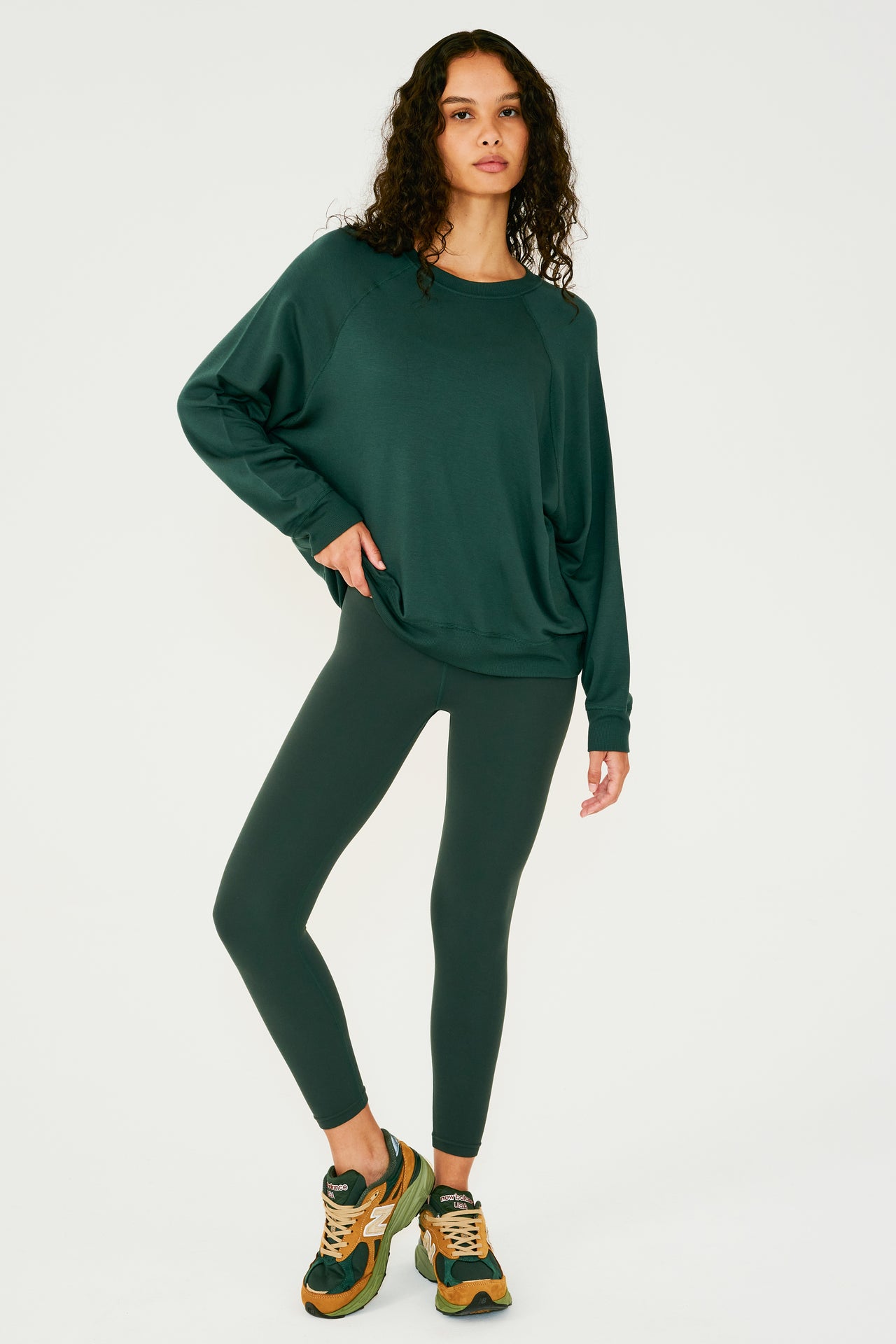 Full front view of girl wearing dark green sweatshirt with visible stitching and ribbed hem, with dark green leggings and multi colored shoes