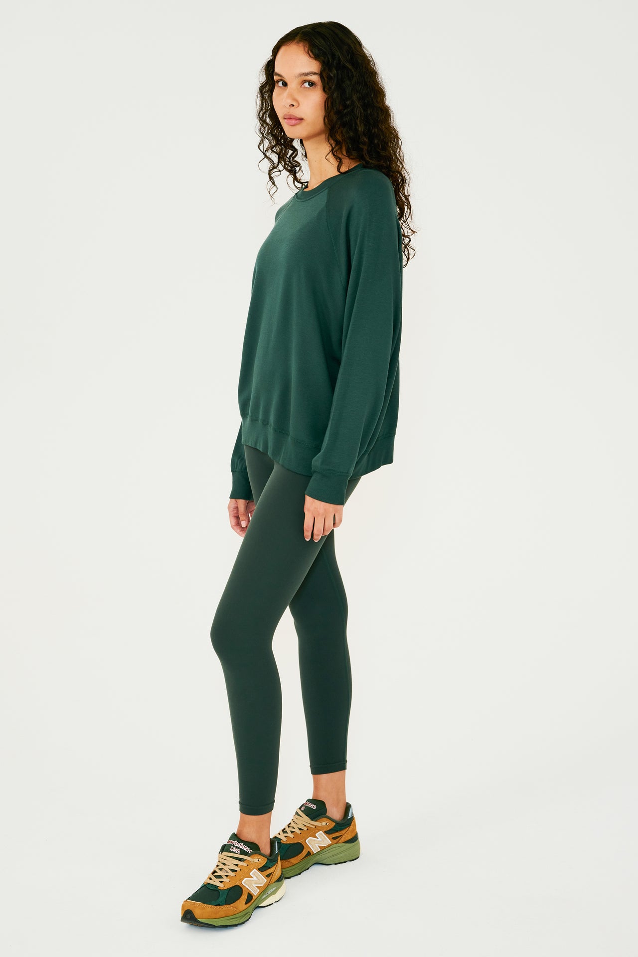 Full side view of girl wearing dark green sweatshirt with visible stitching and ribbed hem, with dark green leggings and multi colored shoes