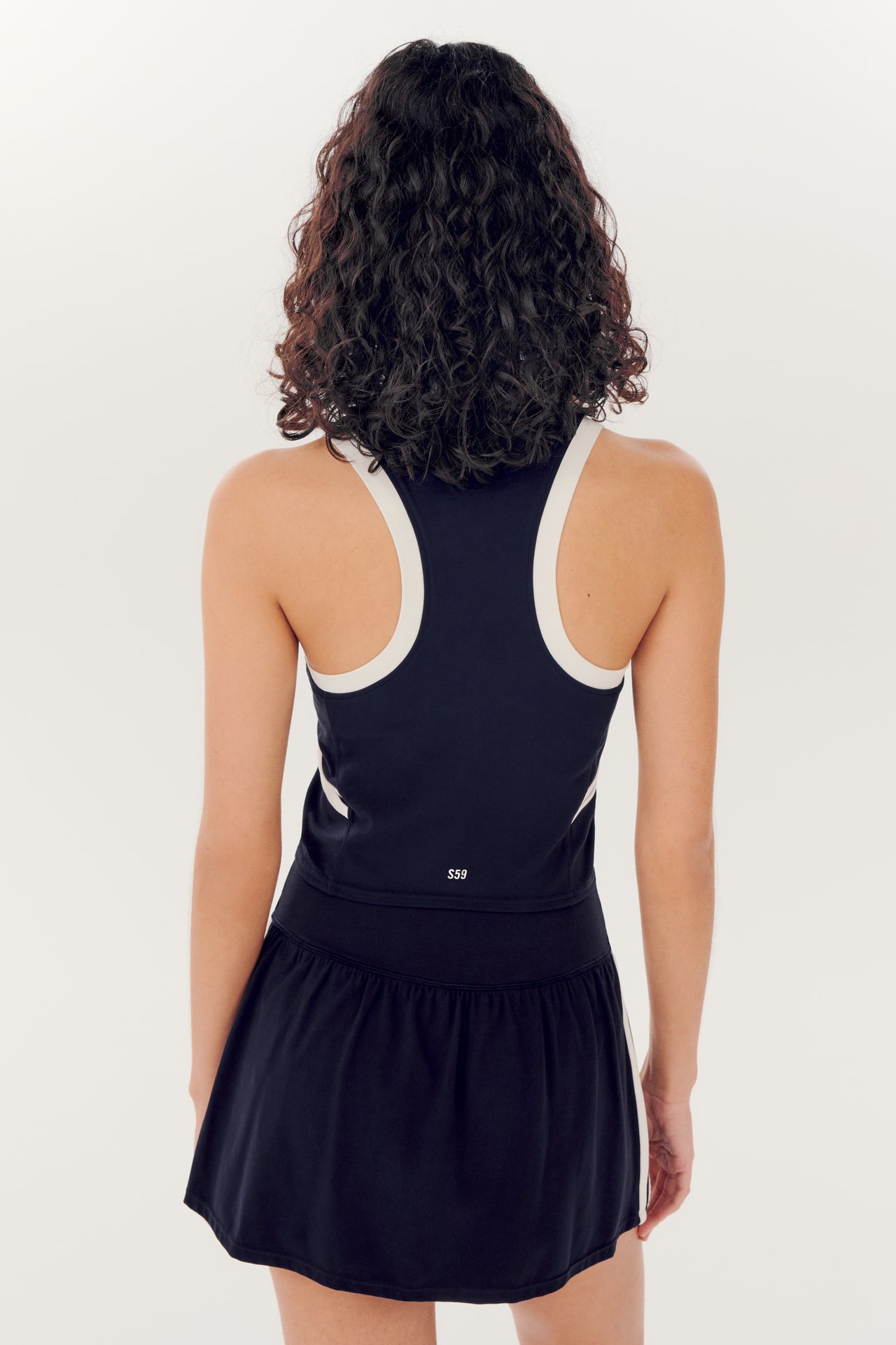 Woman from behind wearing a SPLITS59 Austin Airweight Crop Polo in Indigo/White, made of nylon and spandex fabric, and a black skort.