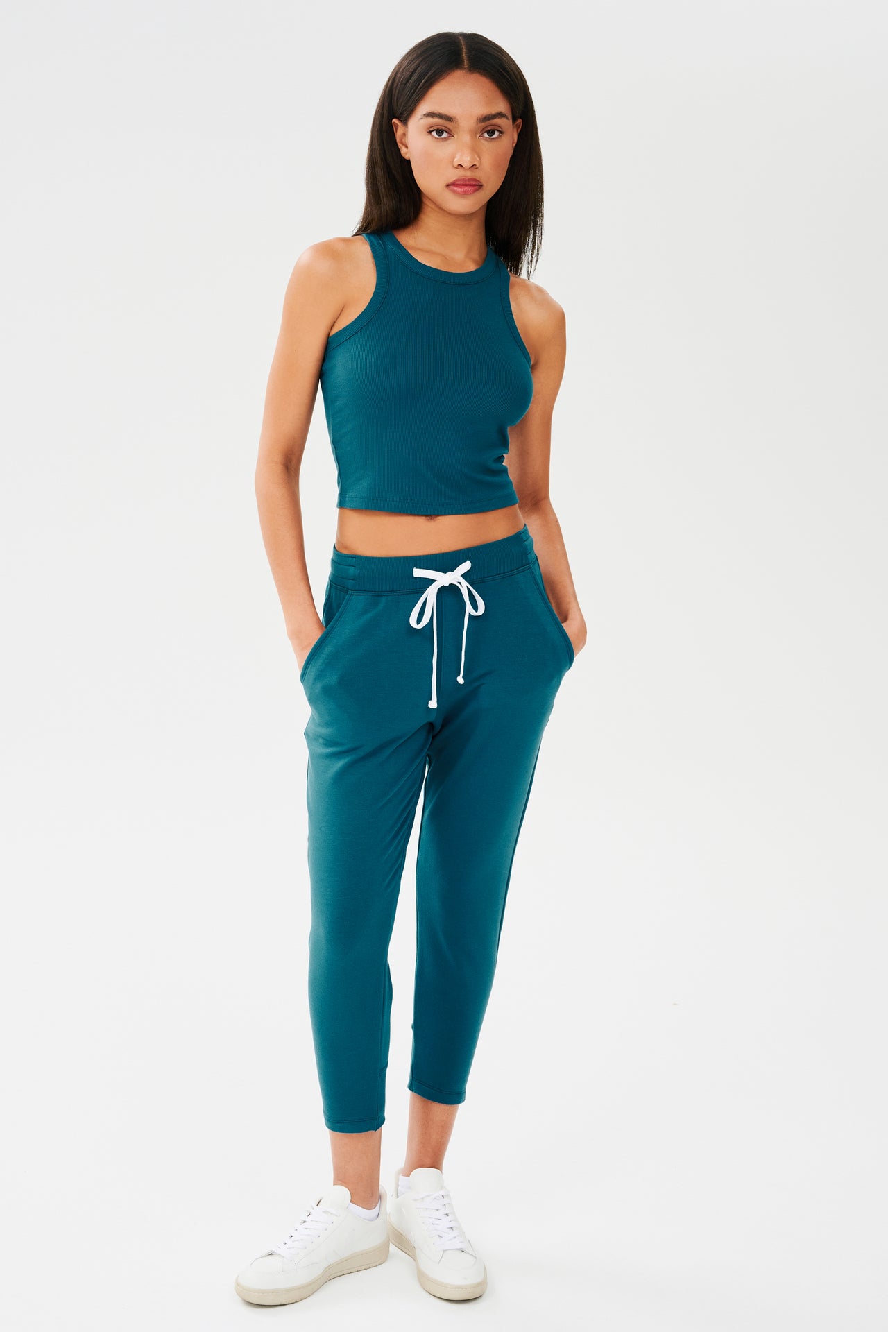 Full front view of woman with dark straight hair wearing a green and blue tone sweatpant with tapered leg and above ankle length with white drawstring and side hip pockets and a green and blue tone high neck ribbed racer front and back tank top. Paired with white shoes. 