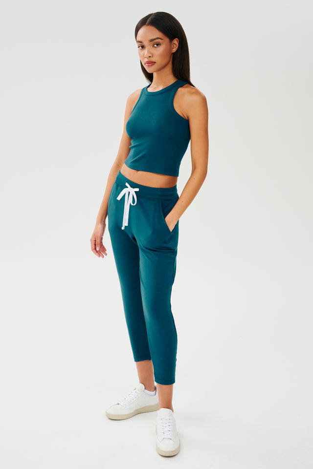 Full front side view of woman with dark straight hair wearing a green and blue tone sweatpant with tapered leg and above ankle length with white drawstring and hip side pickets and a green and blue tone high neck ribbed racer front and back tank top. Paired with white shoes. 