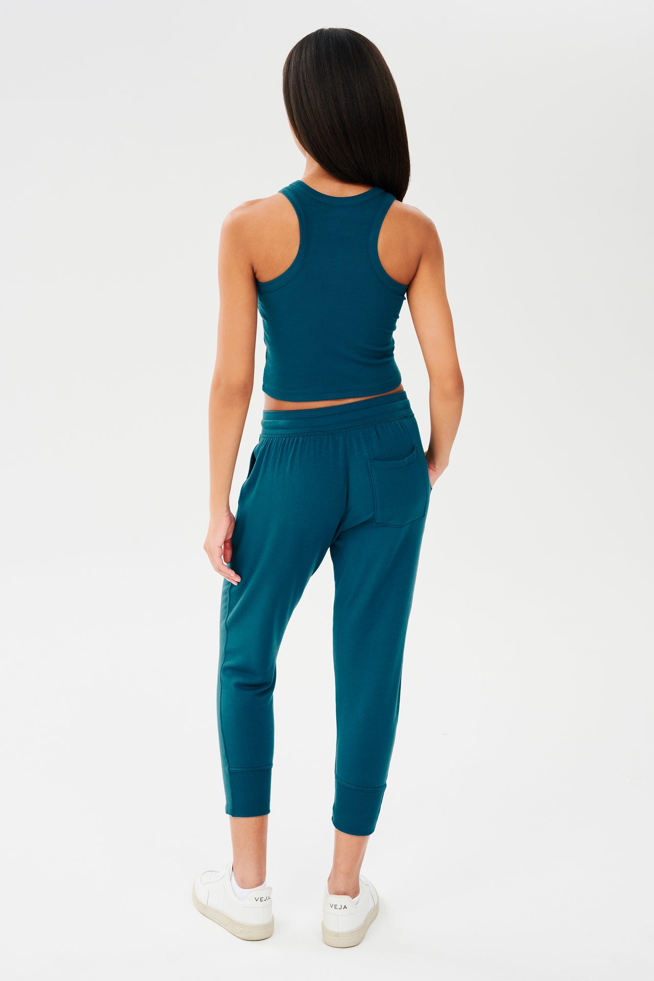 The back view of a woman wearing teal joggers and a SPLITS59 Kiki Rib Crop Tank in Peacock at the gym.