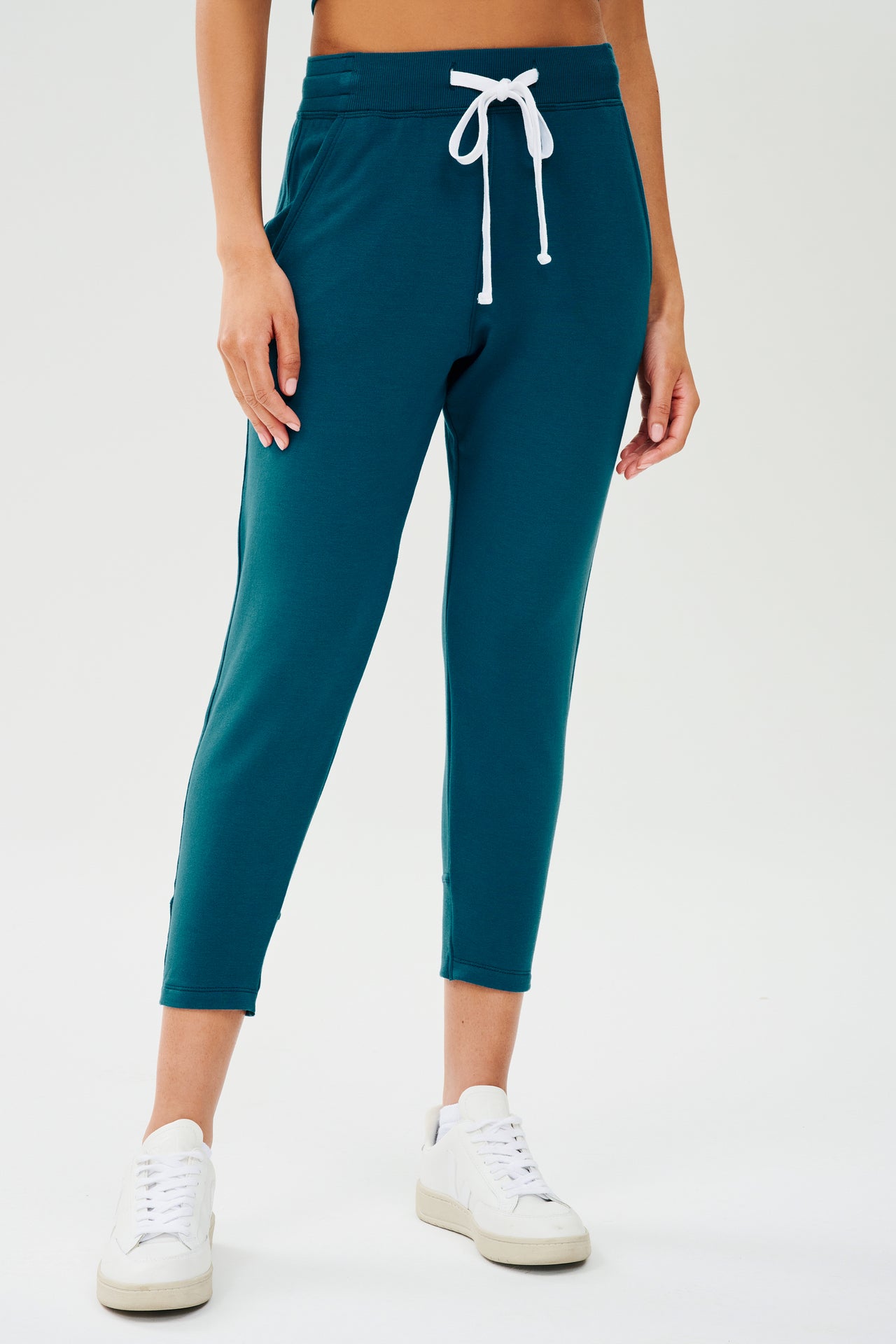 Front view of woman wearing a green and blue tone sweatpant with tapered leg and above ankle length with white drawstring and side hip pockets. Paired with white shoes. 