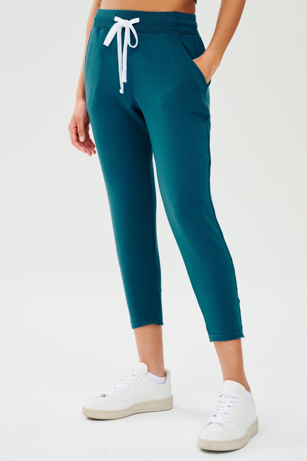 Front side view of woman wearing a green and blue tone sweatpant with tapered leg and above ankle length with white drawstring and side hip pockets. Paired with white shoes.