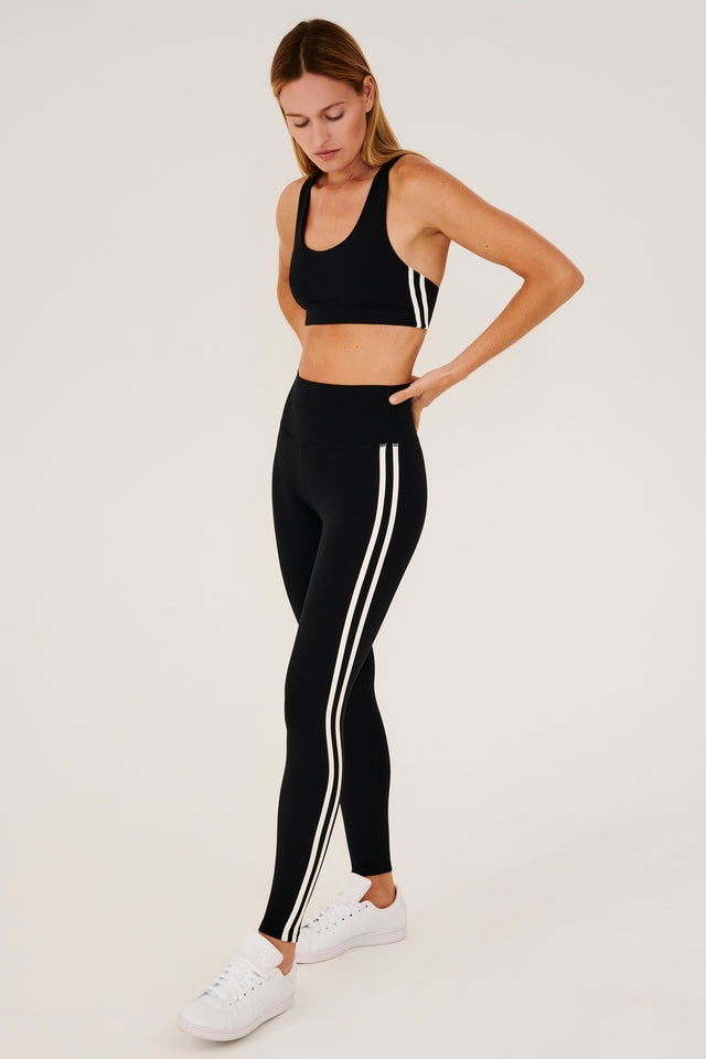 Full side view of girl wearing black leggings with two thin white stripes down the side and a black sports bra with white shoes