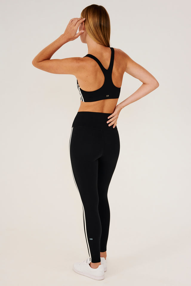 Full back view of girl wearing black sports bra with two thin white stripes  down the side and black leggings with white shoes