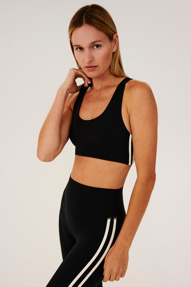 Side view of girl wearing black sports bra with two thin white stripes  down the side and black leggings 