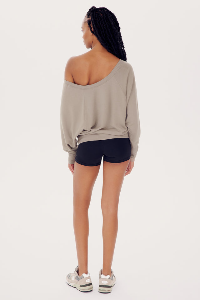 A woman stands facing away from the camera, wearing a SPLITS59 Indy Dolman Fleece Sweatshirt in Latte and black shorts, with her hair styled in long braids.