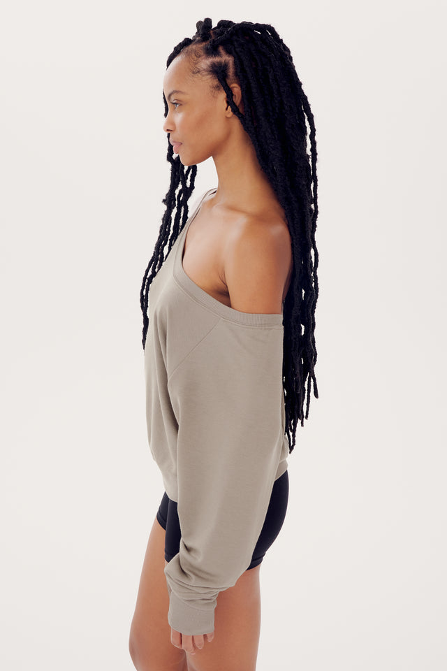 Woman with long braided hair wearing a SPLITS59 Indy Dolman Fleece Sweatshirt in Latte and black shorts, standing in profile against a white background.