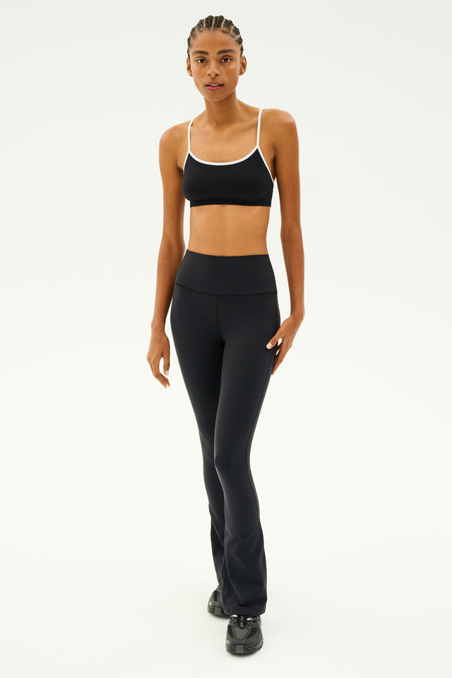 Dressed in the SPLITS59 Loren Seamless Bra in Black/White, paired with chafe-free leggings and black sneakers, a person stands poised in a studio, ready for intense gym workouts.