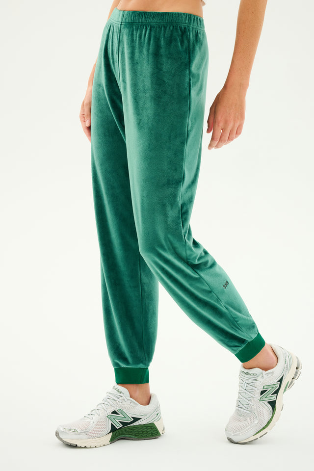 A person lounging in Rhys Velour Jogger pants in the Arugula color by SPLITS59 and white New Balance sneakers stands against a light background.