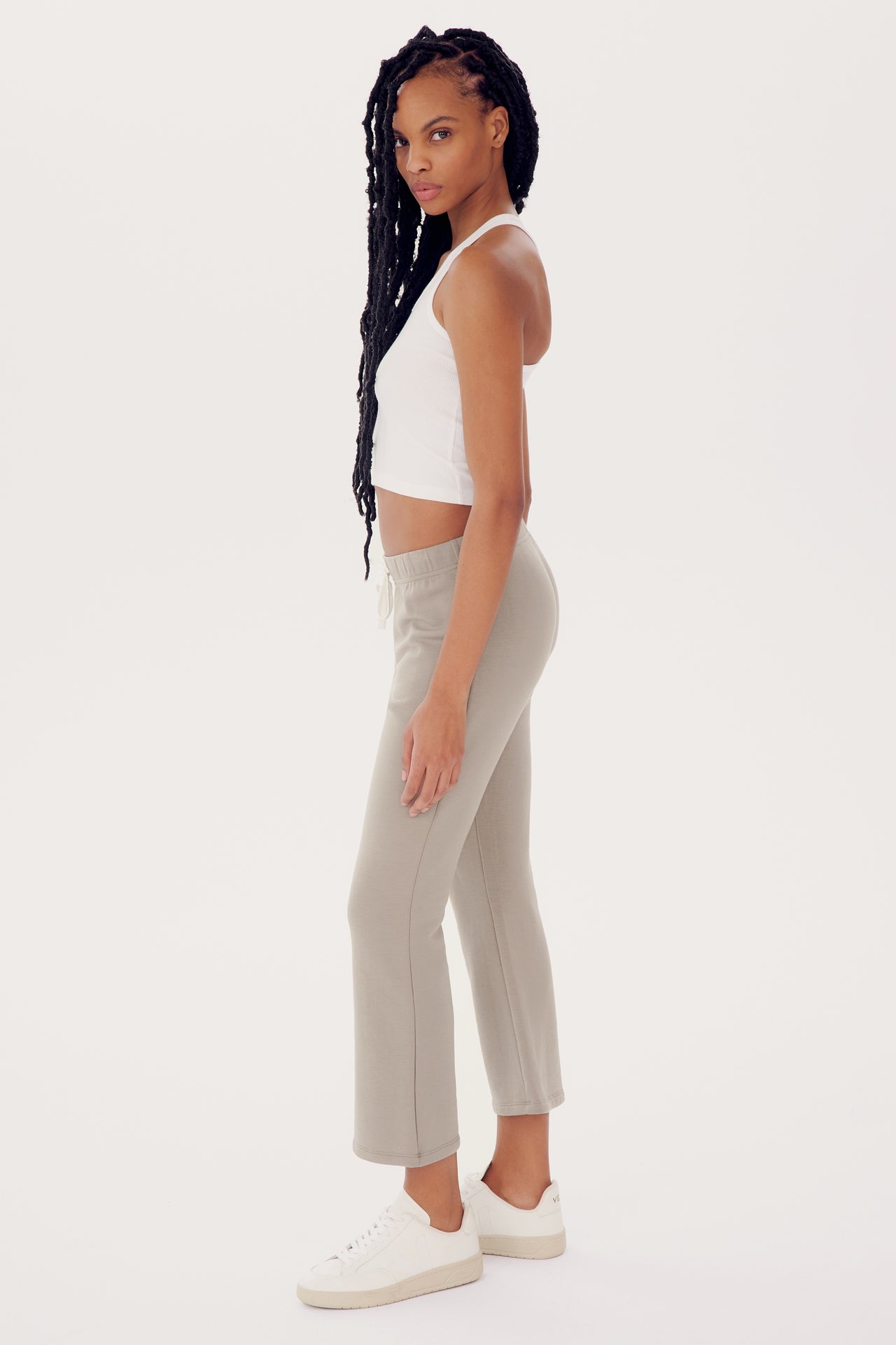 A woman in a white tank top and SPLITS59 beige flared trousers stands sideways, looking over her shoulder, with a neutral expression and long braided hair.