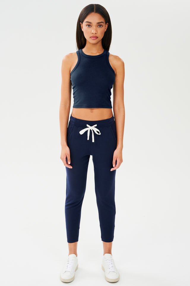 Full front view of woman with dark straight hair wearing a dark  blue sweatpant with tapered leg and above ankle length with white drawstring and side hip pockets and a dark blue high neck ribbed racer front and back tank top. Paired with white shoes.