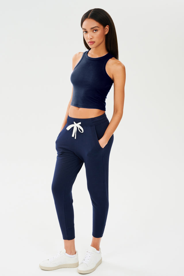 Full front side view of woman with dark straight hair wearing a dark  blue sweatpant with tapered leg and above ankle length with white drawstring and side hip pockets and a dark blue high neck ribbed racer front and back tank top. Paired with white shoes.