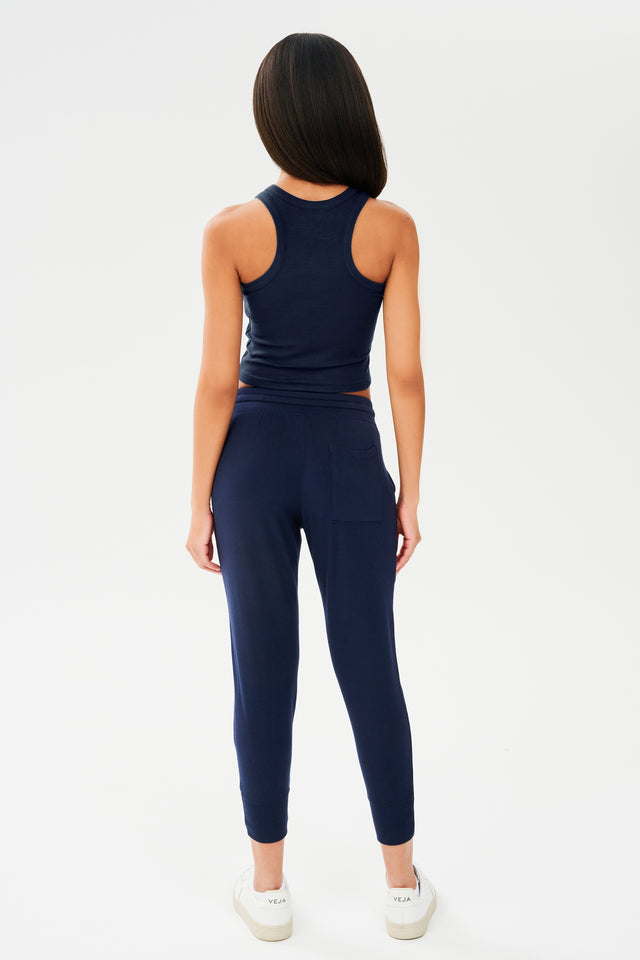 Full back view of woman with dark straight hair wearing a dark blue sweatpant with tapered leg and above ankle length with side hip pockets and a dark blue high neck ribbed racer front and back tank top. Paired with white shoes.