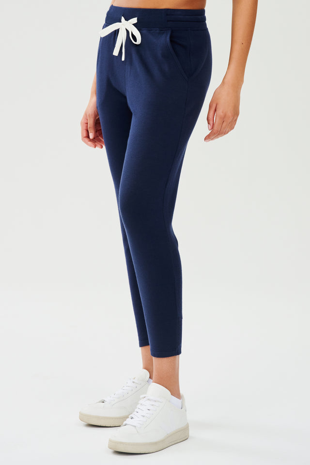 Front side view of woman wearing a dark blue sweatpant with tapered leg and above ankle length with white drawstring and side hip pockets. Paired with white shoes.