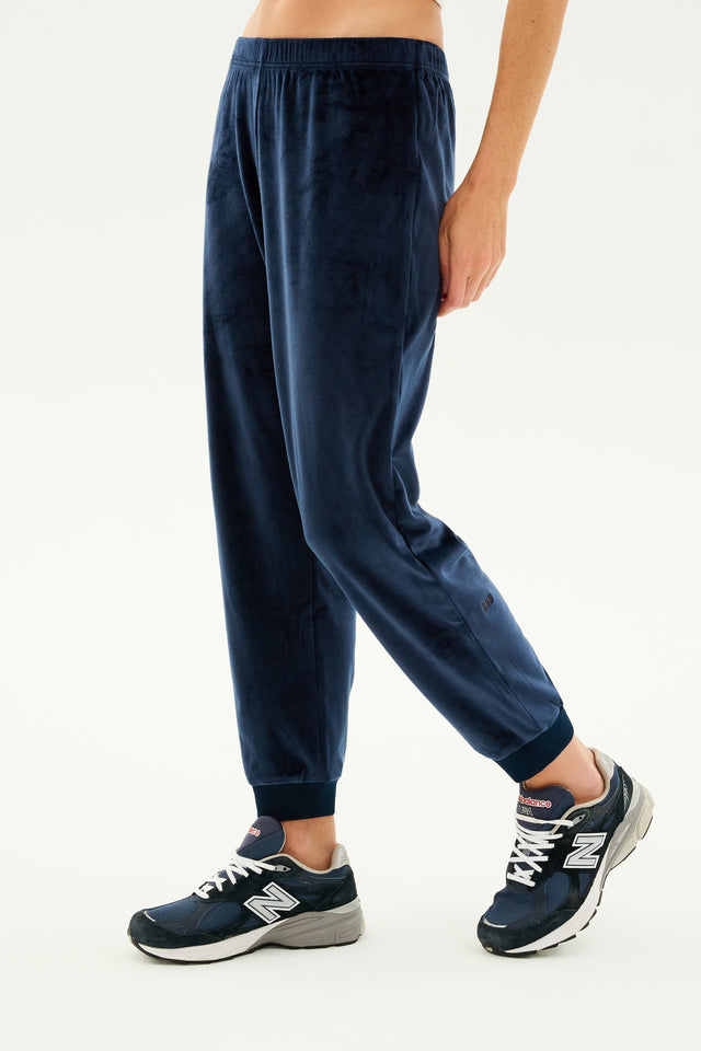 Person lounging in Rhys Velour Jogger - Indigo from SPLITS59 and blue New Balance sneakers. The image focuses on the lower part of their body against a plain, light-colored background, showcasing casual wear perfect for relaxation.